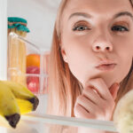 Frau blickt nachdenklich in einen Kühlschrank voller Obst und Gemüse.