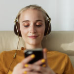 Frau in einem gelben Oberteil entspannt auf einer Couch, trägt Kopfhörer und schaut auf ein Smartphone.