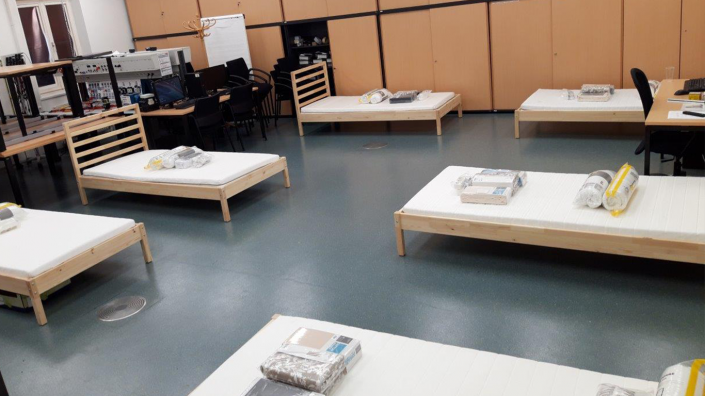 Zimmer mit sechs Betten, jeweils mit Bettzeugpaketen, im Hintergrund Schreibtische mit Stühlen.