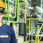 Ein mit Augmented-Reality-Brille und Schutzkleidung ausgestatteter Arbeiter steht in einer Industrieanlage, umgeben von grünen Rohren und Sicherheitsgeländern.
