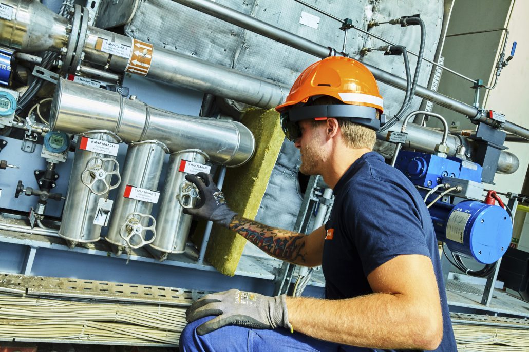 Arbeiter in Sicherheitsausrüstung überprüft Maschinen mit Messgeräten und Rohren in einer industriellen Umgebung und trägt dabei eine Datenbrille.