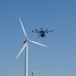 Eine Drohne fliegt vor einem klaren blauen Himmel in der Nähe von einer Windturbine.