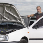 Werner Beck stand neben einem weißen E-Auto mit geöffneter Motorhaube, umgeben von der weiten Natur unter einem bewölkten Himmel. Das Auto hat er selbst gebaut.