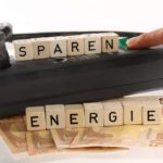 Auf einer Steckdosenleiste sind Holzklötze mit der Aufschrift „SPAREN ENERGIE“ zu sehen, darunter Euroscheine und ein Finger, der einen Schalter drückt.