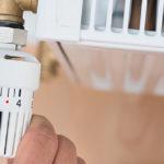 Hand stellt den Temperaturregler am Thermostatventil eines weißen Heizkörpers ein.