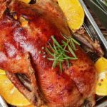 Gebratene Ente, garniert mit Rosmarin und umgeben von Orangenhälften in einer Metallauflaufform.