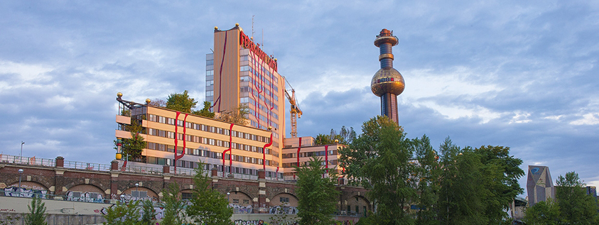 Farbenfrohe Gebäude mit einem runden Turm in einer üppigen Grünanlage unter einem bewölkten Himmel.