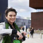 Elisabeth Oberzaucher lächelt mit kurzen dunklen Haaren und einem grünen Schal, der elegant um ihren Hals drapiert ist. Sie steht im Freien vor dem Viertel Zwei.