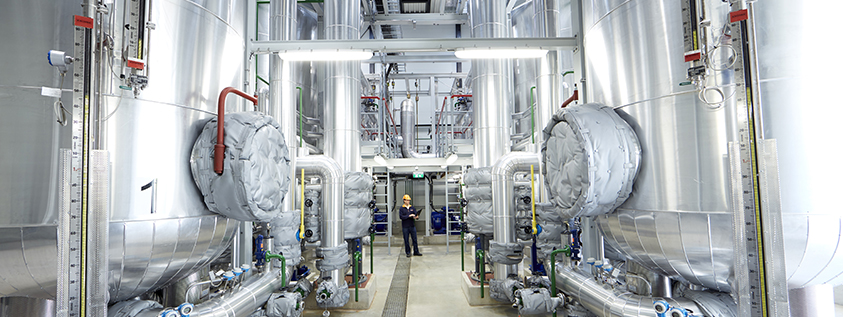 Industrieanlage mit großen silbernen Tanks und Rohren. - power-2-heat-Anlage In der Mitte steht eine Person mit Schutzhelm.