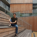 Eine Person sitzt auf Holzstufen in einem modernen Innenhof mit holzgetäfelten Gebäuden im Hintergrund.