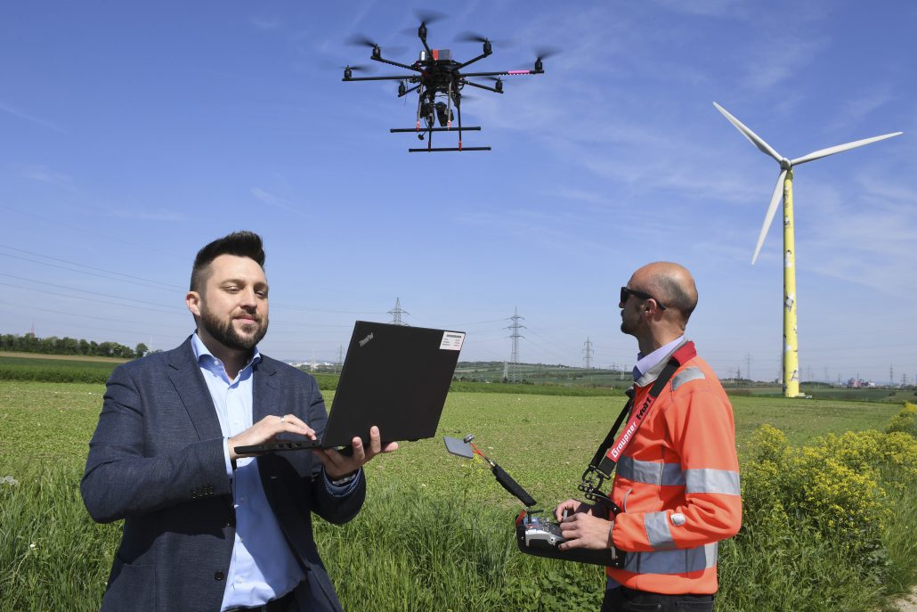 Zwei Männer bedienen eine Drohne in der Nähe einer Windturbine auf einer Wiese. Einer verwendet einen Laptop, der andere hält eine Drohnen-Steuerung in der Hand.