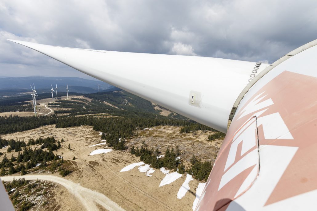 Vom Standpunkt eines Windradflügels im Windpark Steinriegel aus betrachtet, ragen in der Ferne stolze Turbinen unter einem wolkigen Himmel auf, vor der Kulisse einer majestätischen Berglandschaft.