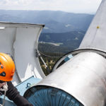 Ein Arbeiter mit einem orangefarbenen Helm führt Wartungsarbeiten an einer Windkraftanlage mit Bergen im Hintergrund durch.