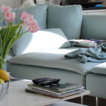 Ein helles Wohnzimmer verfügt über ein hellblaues Sofa, rosa Tulpen in einer Vase und eine Schale mit gemischtem Obst auf einem weißen Couchtisch. Zeitschriften und eine Fernbedienung liegen ordentlich auf dem Tisch.