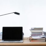 Laptop, Schreibtischlampe, Bücher und Ordner auf einem Holzschreibtisch vor einem schlichten weißen Hintergrund.