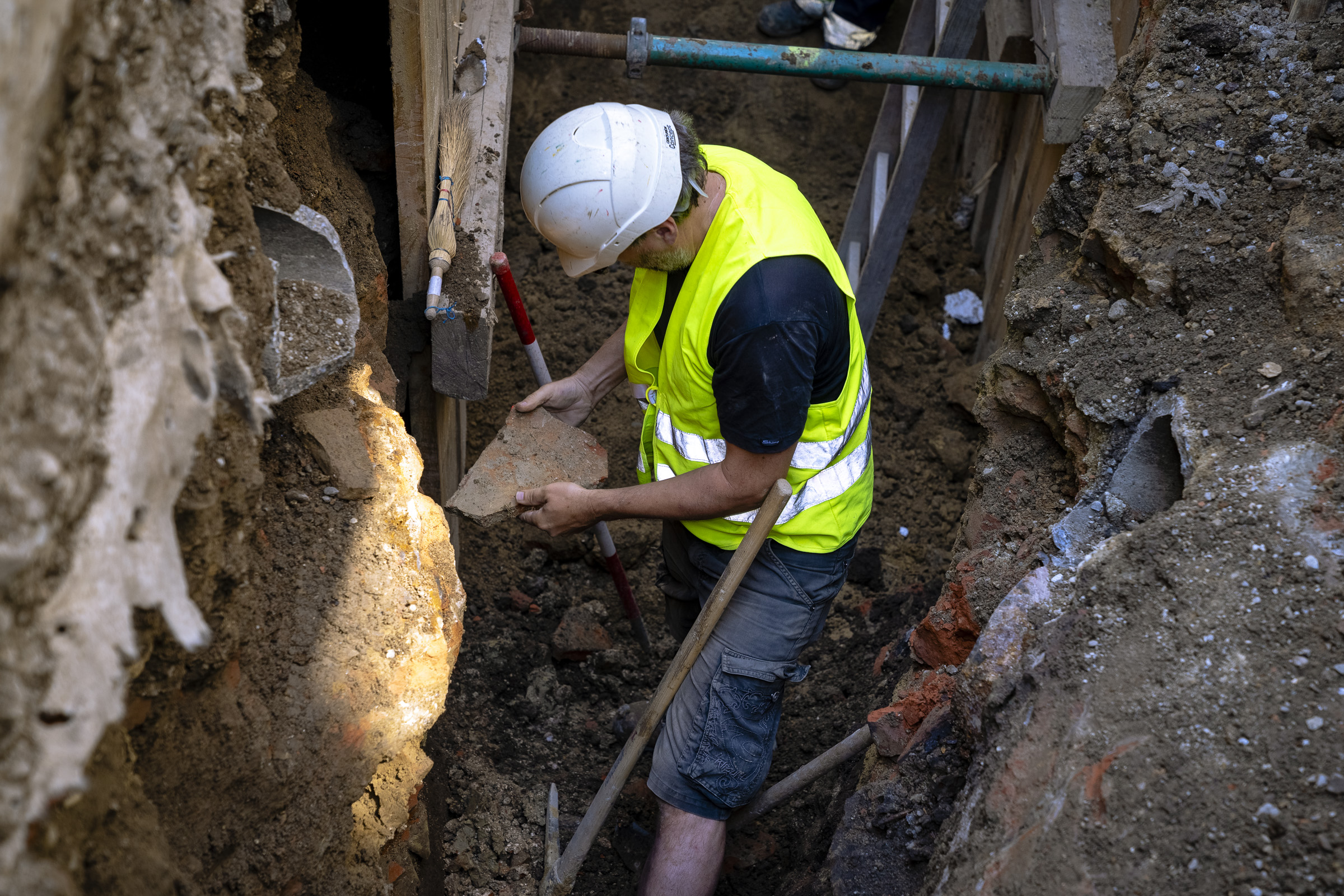 Bauarbeiter untersucht in einem Graben ein historisches Felsfragment, während er einen Schutzhelm und eine Warnweste trägt.