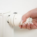 Hand, die einen weißen Heizkörperthermostat einstellt, trägt einen gelben Pullover und ein silbernes Armband.