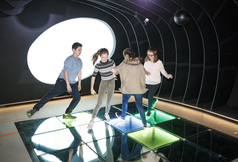 Vier Kinder tanzen fröhlich auf einem Glasboden, der von bunten Lichtern in einem dunklen Raum beleuchtet wird. Die Szene ist vor dem Hintergrund einer großen ovalen Leuchte inszeniert und fängt die lebendige Energie ein, die an die Erlebniswelt Spittelau erinnert.