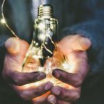 Hände halten eine leuchtende Glühbirne voller kleiner Lichterketten vor einem dunklen Hintergrund.