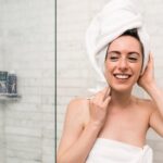 Eine lächelnde Frau mit Handtuch und umwickeltem Kopf steht in einer Dusche und berührt freudig ihr Gesicht.