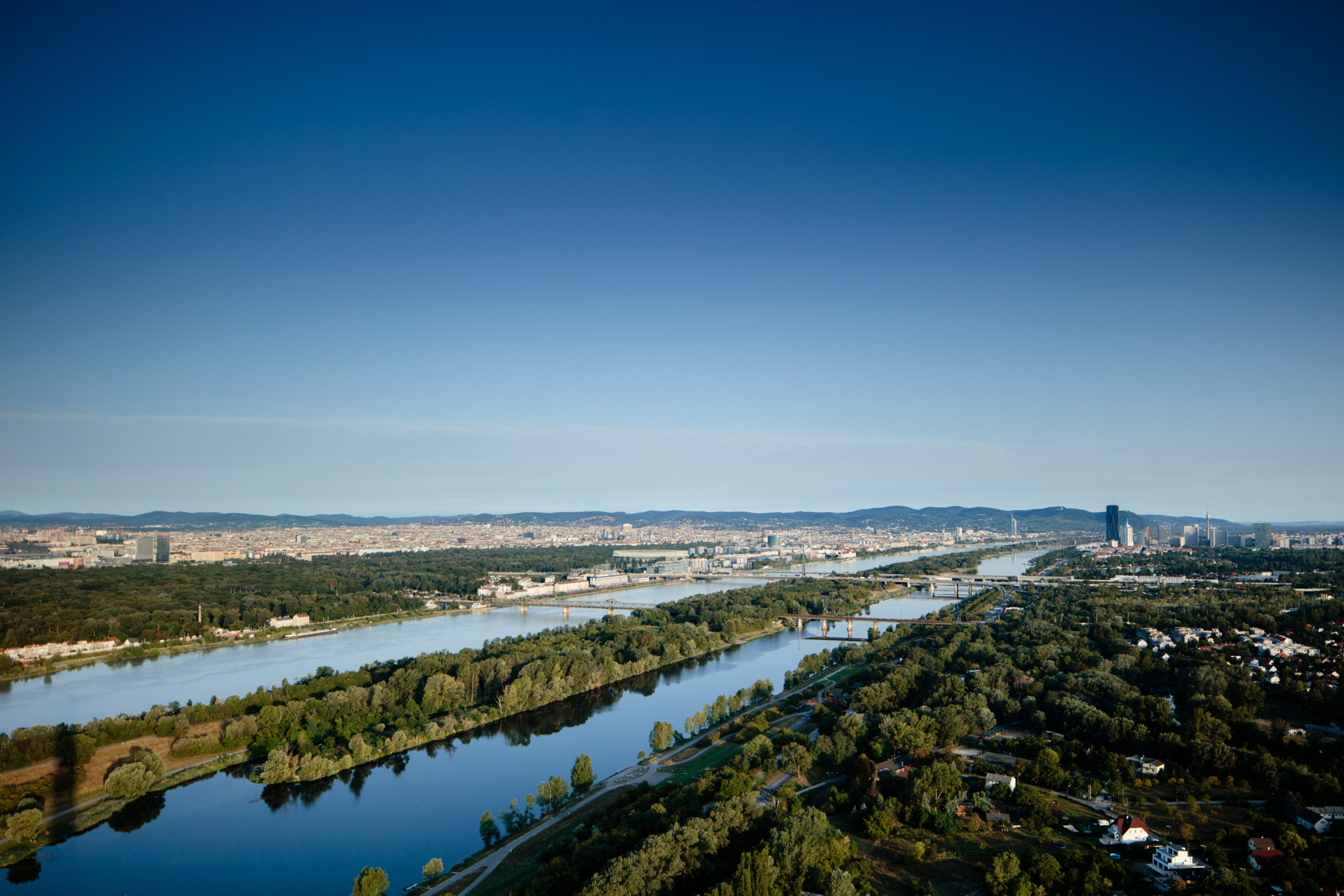 Luftaufnahme der Wiener Stadtlandschaft mit der Donau, grünen Parks und Bergen in der Ferne unter einem klaren blauen Himmel.