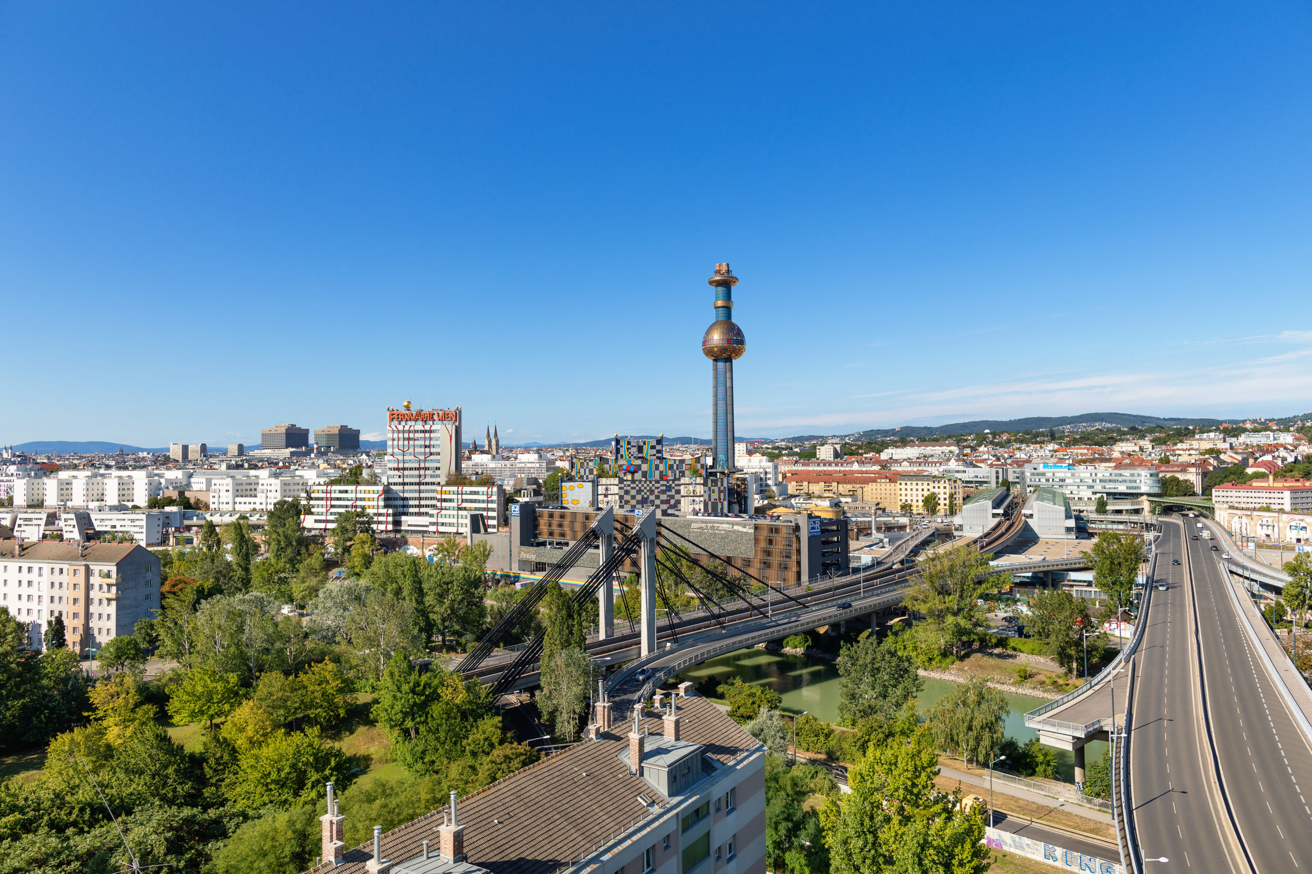 Luftaufnahme der Wiener Stadtlandschaft mit dem ikonischen Turm der Abfallverwertungsanlage Spittelau, umgeben von Gebäuden, Grünflächen und einer Autobahn unter dem klaren blauen Himmel.