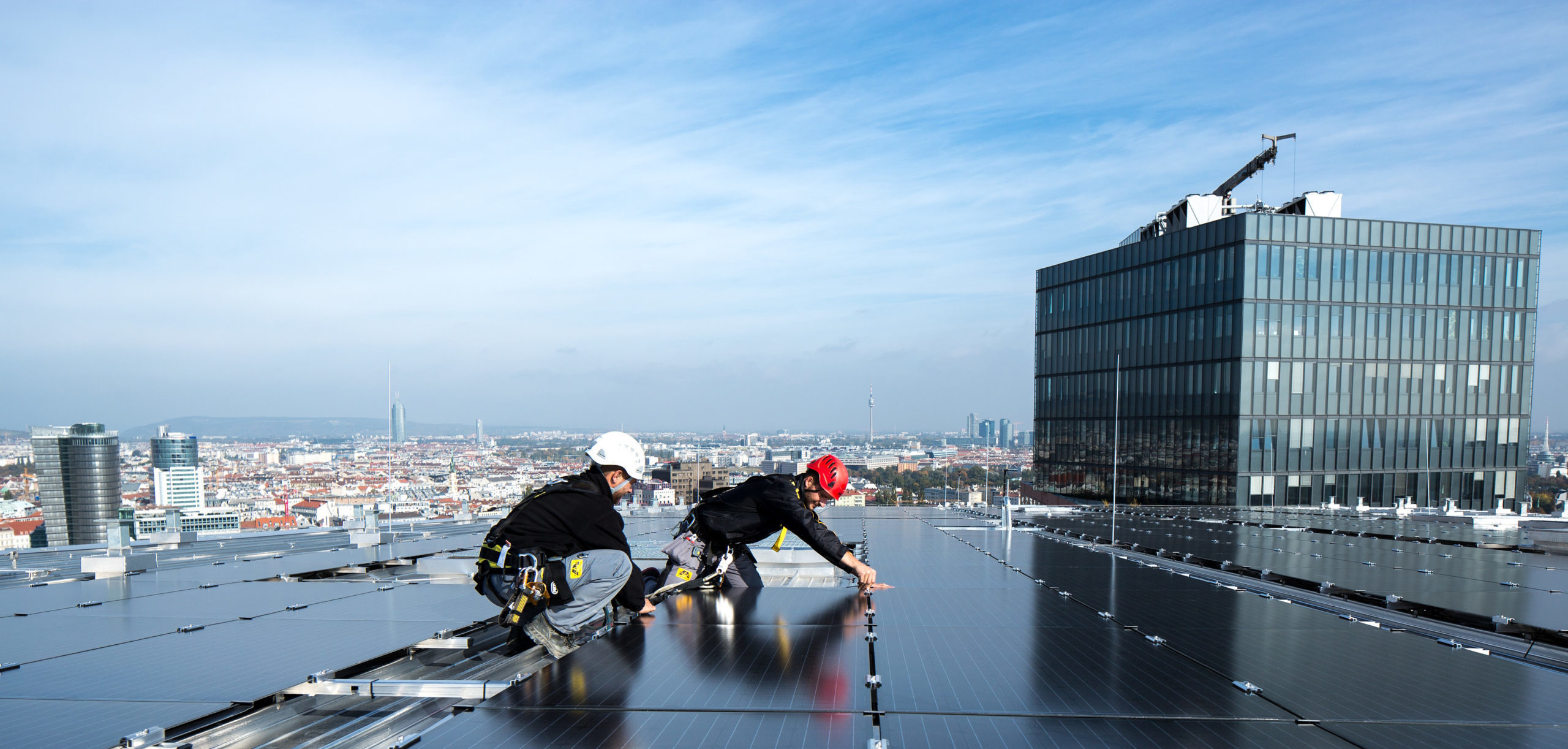Zwei Arbeiter in Sicherheitsausrüstung installieren Solarmodule der Photovolaikanlage Wien Mitte auf dem Dach. Im Hintergrund ist die Stadt Wien zu sehen.