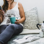 Frau entspannt auf einem Sofa mit Telefon und Tasse in der Hand, auf dem Tisch steht ein offenes Buch und eine Kaffeekanne.