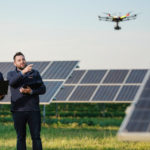 Zwei Wien Energie Mitarbeiter beobachten mit Laptops eine Drohne, die in der Nähe von Solarmodulen auf einer Wiese fliegt, und sorgen so für eine präzise Drohnenwartung.