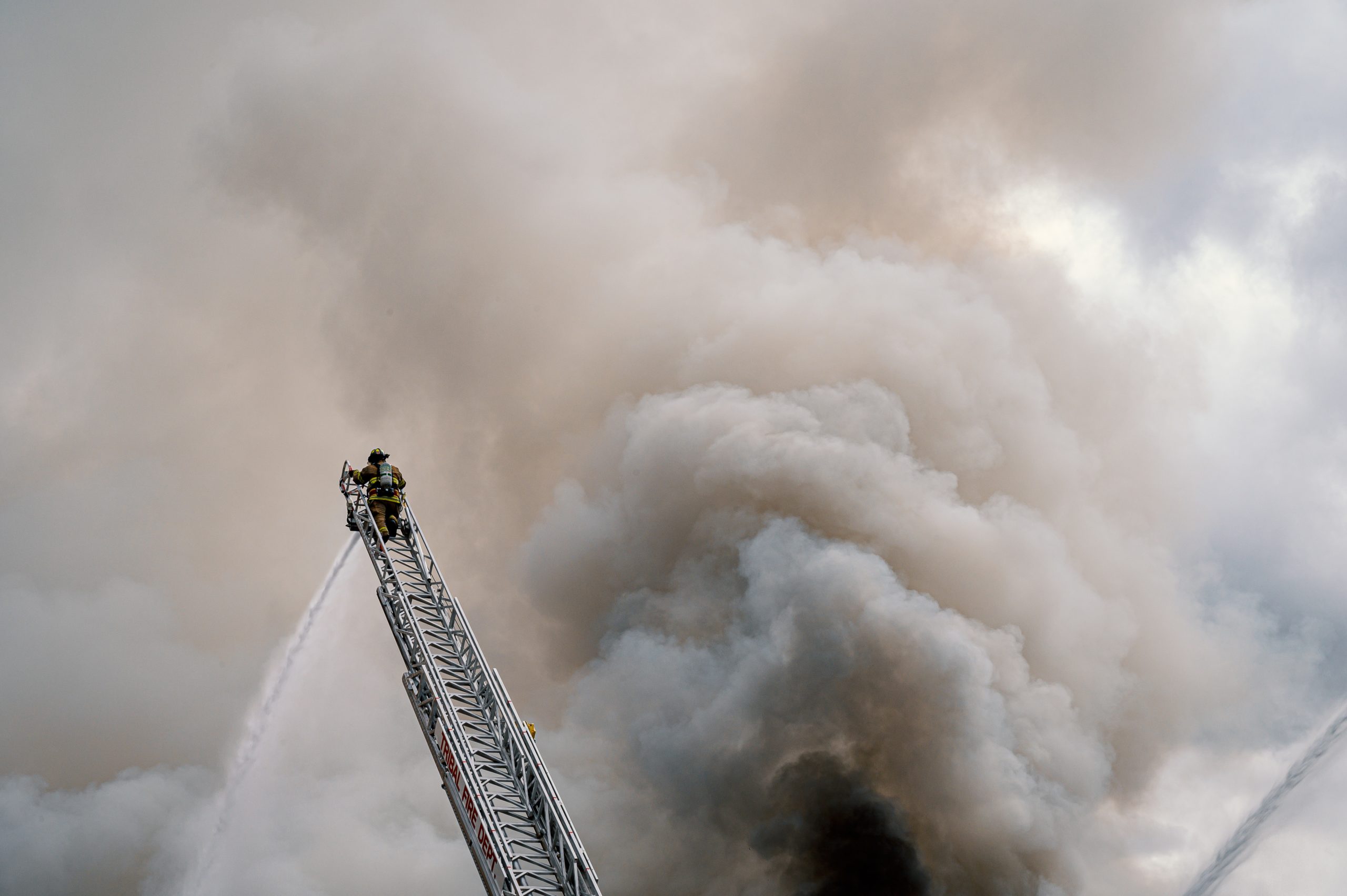 Feuerwehrmann auf einer Leiter sprüht Wasser auf eine große Rauchwolke.