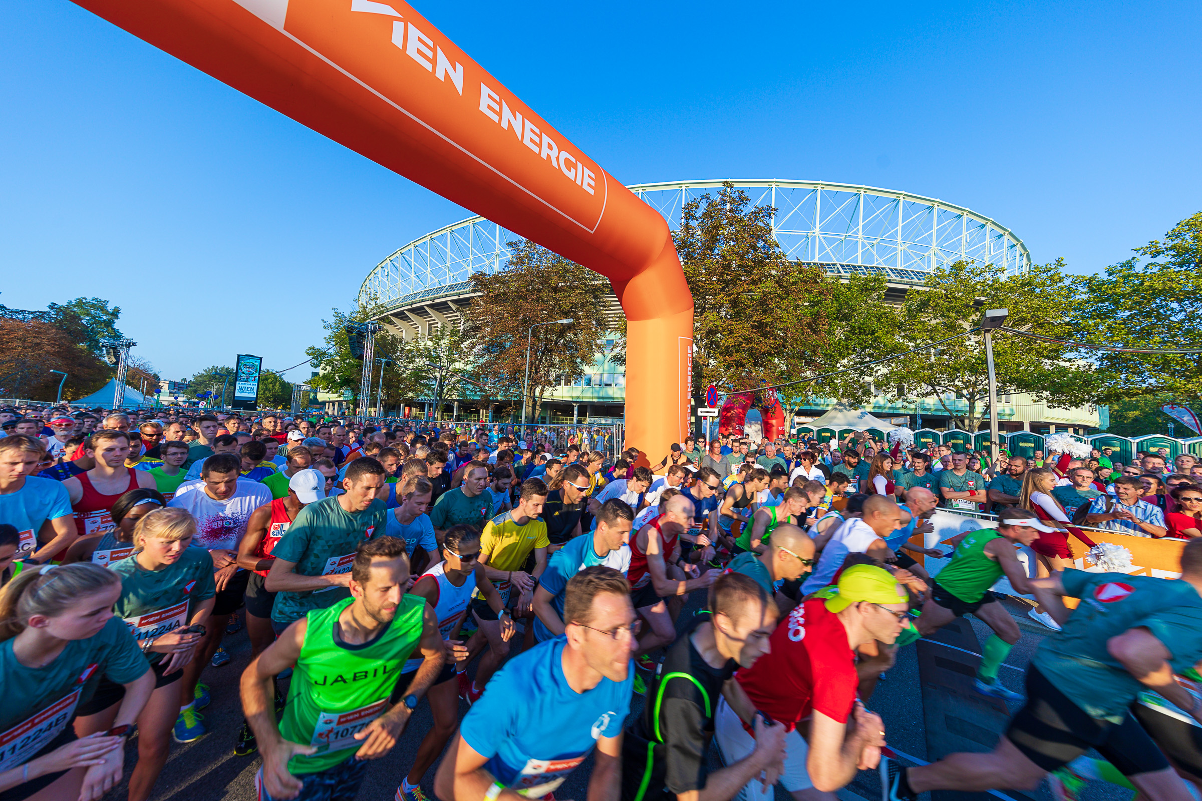 Eine Menge Läufer starten unter einem großen orangefarbenen Bogen mit der Aufschrift "Wien Energie" ein Rennen. Im Hintergrund ist ein Stadion zu sehen.