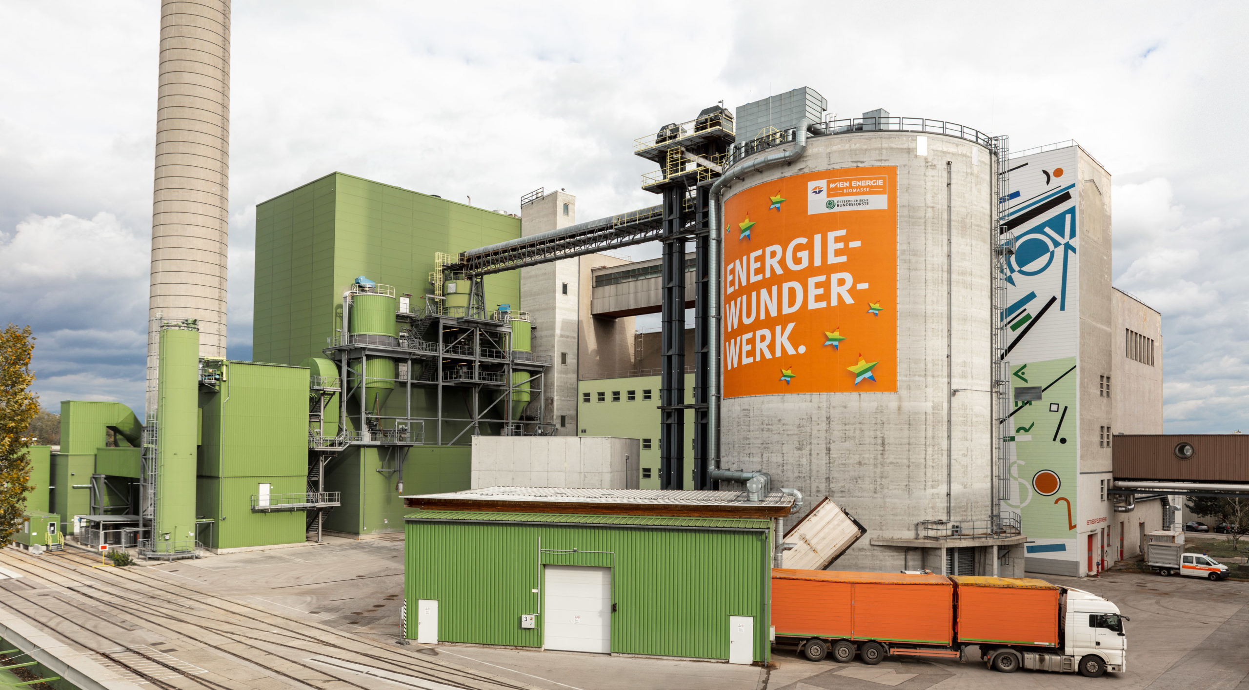 Biomassekraftwerk Simmering mit grünen Gebäuden und einem zylindrischen Turm mit der Aufschrift "Energiewunderkraftwerk".