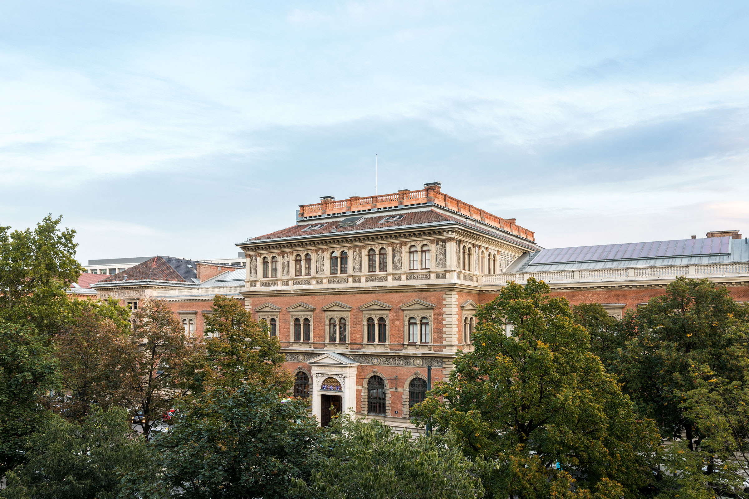 Eingebettet in einen strahlend blauen Himmel ist das historische Backsteingebäude des MAK am Stubenring anmutig von üppigen Bäumen umgeben. Das MAK am Stubenring wird mit Fernkälte von Wien Energie temperiert.