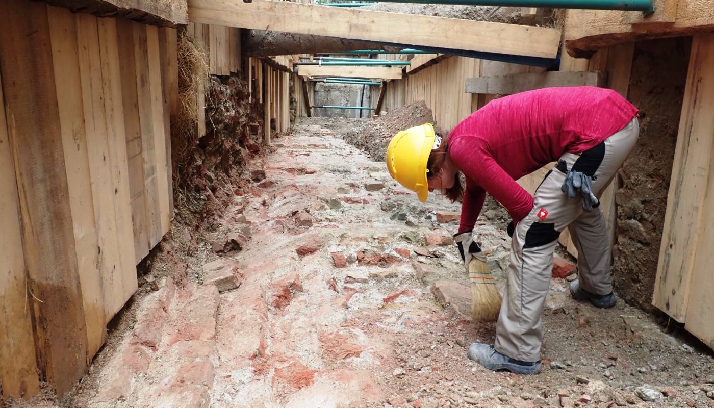 Inmitten einer Fernkälte-Grabung in Wien streift ein Arbeiter in einem Graben, ausgerüstet mit Holzstützen und einem Schutzhelm, vorsichtig den Boden, um Ziegel und Erdschichten freizulegen.