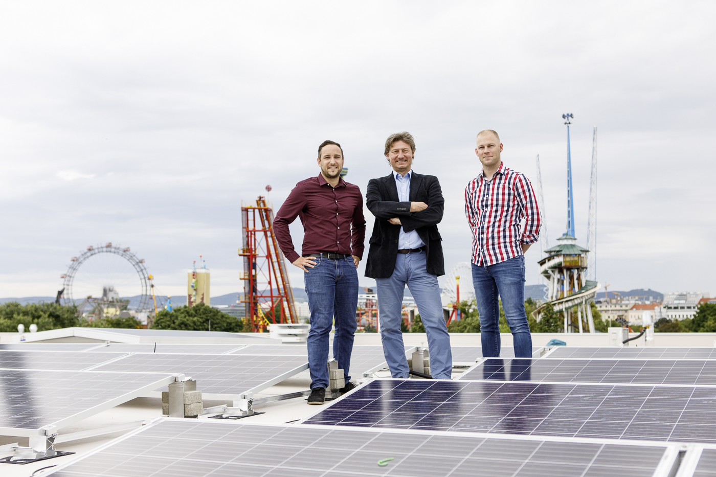 Drei Männer stehen auf einer Photovoltaikanlage auf dem Dach des Königreichs der Eisenbahnen, im Hintergrund leuchtet der Wiener Prater.