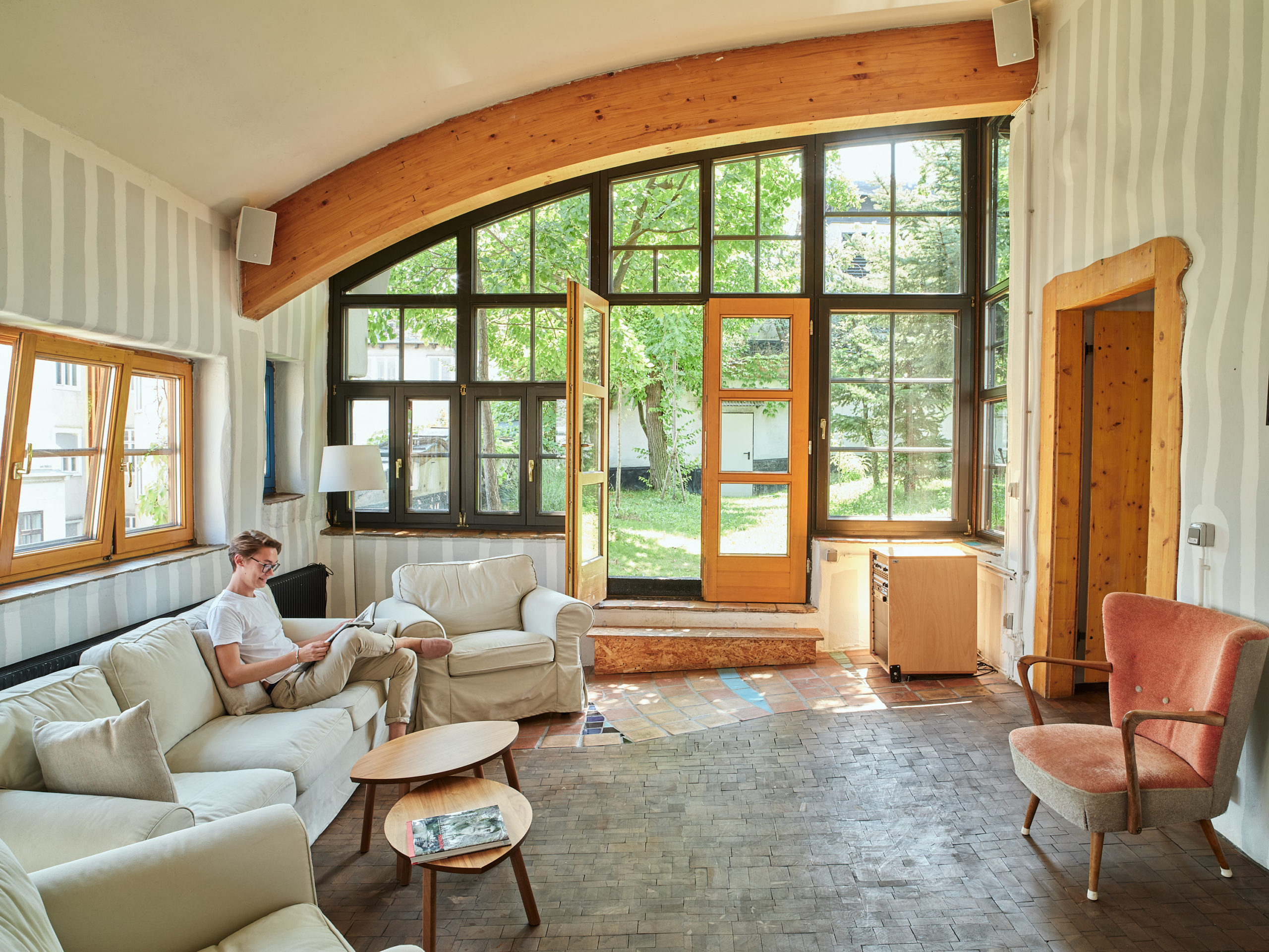 Mann liest in einem sonnendurchfluteten Raum mit großen Fenstern, weißen Sofas, einem hölzernen Couchtisch und freiem Blick auf den Garten. Von Friedensreich Hundertwasser gestaltetes Wohnzimmer im Kunst Haus Wien