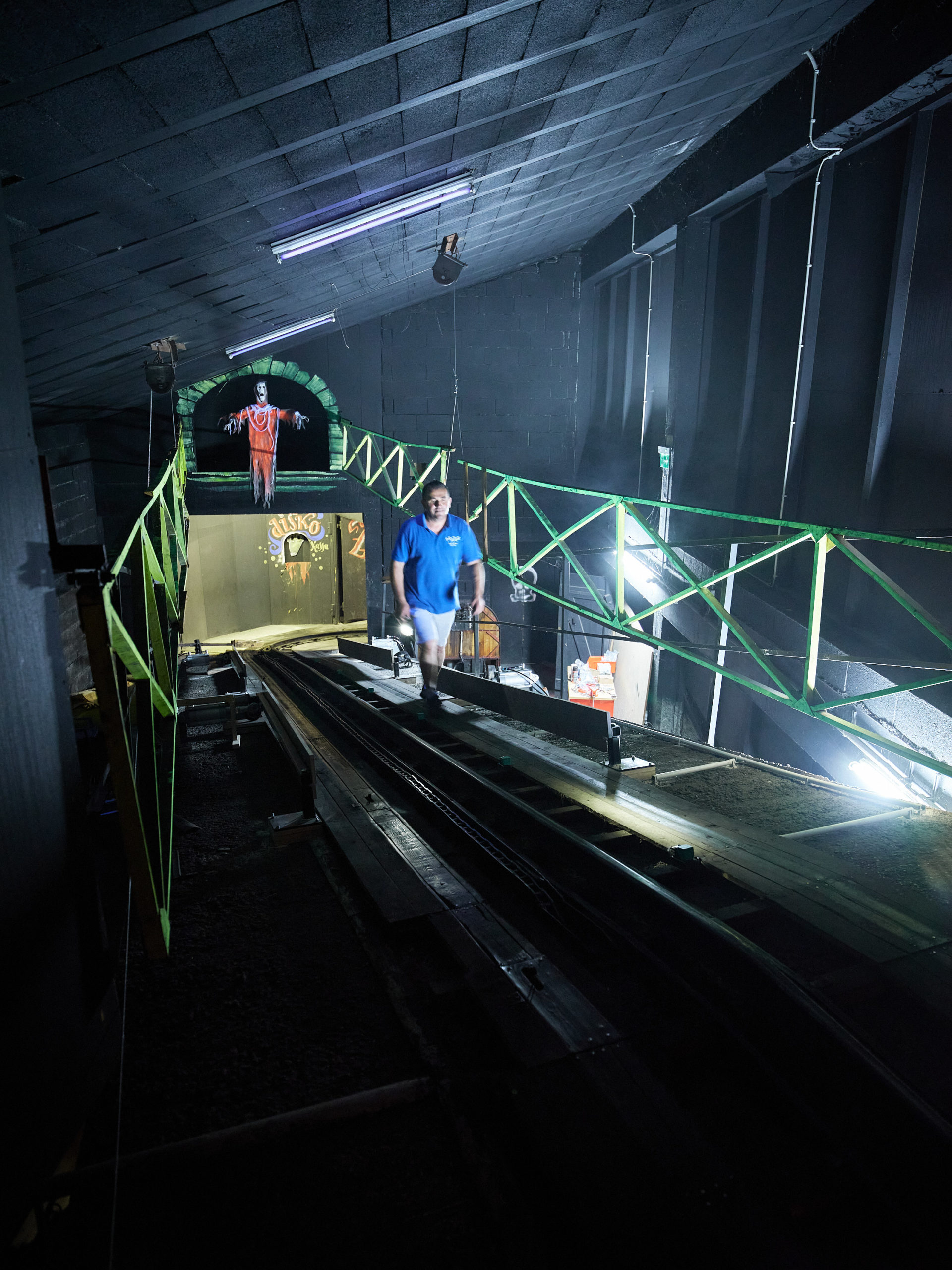Eine Person geht in der Geisterbahn auf einem schwach beleuchteten Weg mit grünem Geländer entlang.