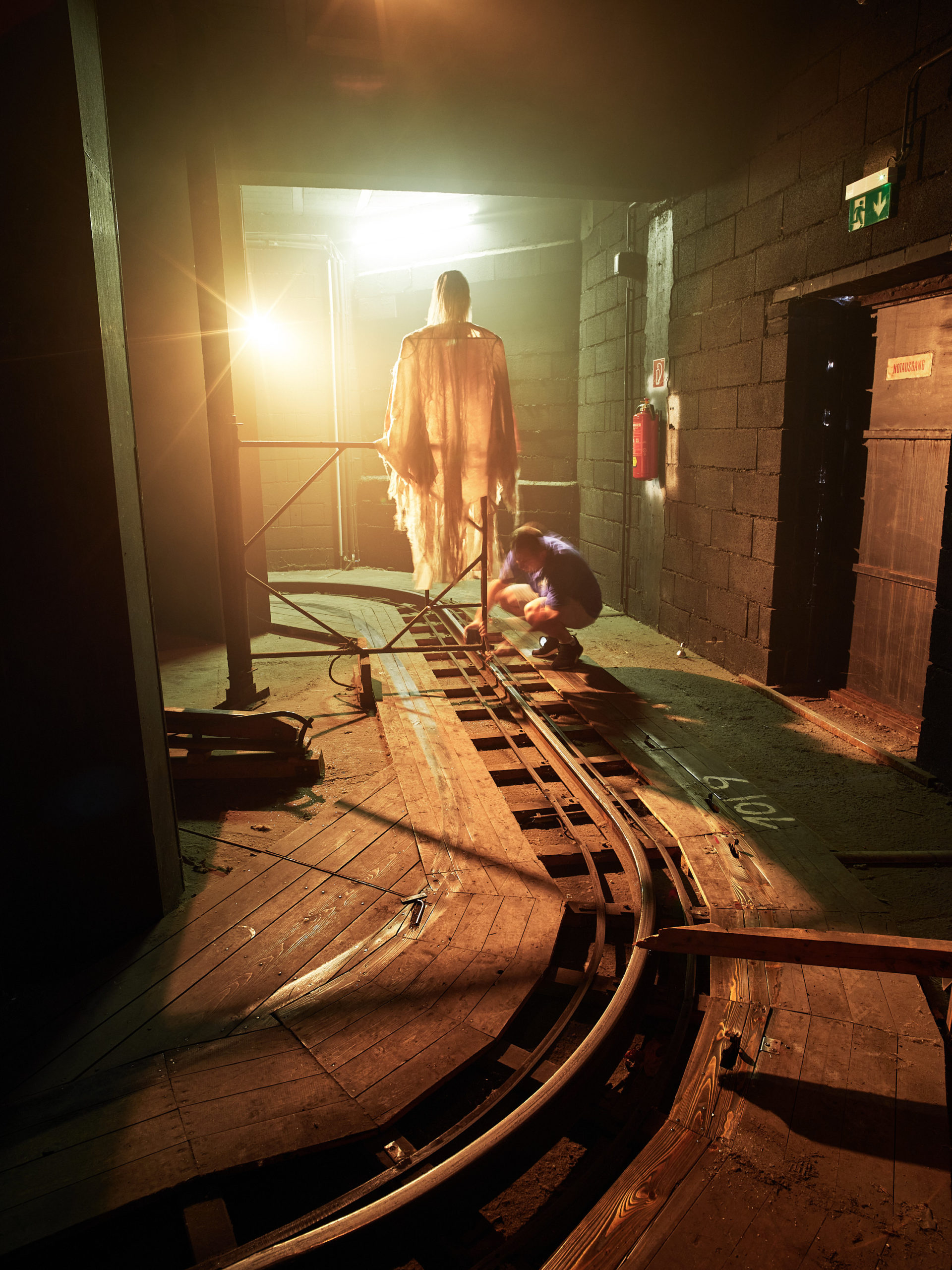 Dunkler Raum der Geisterbahn im Wiener Prater. Person repariert etwas an den Gleisen. Im Hintergrund ist eine Geisterfigur zu sehen.