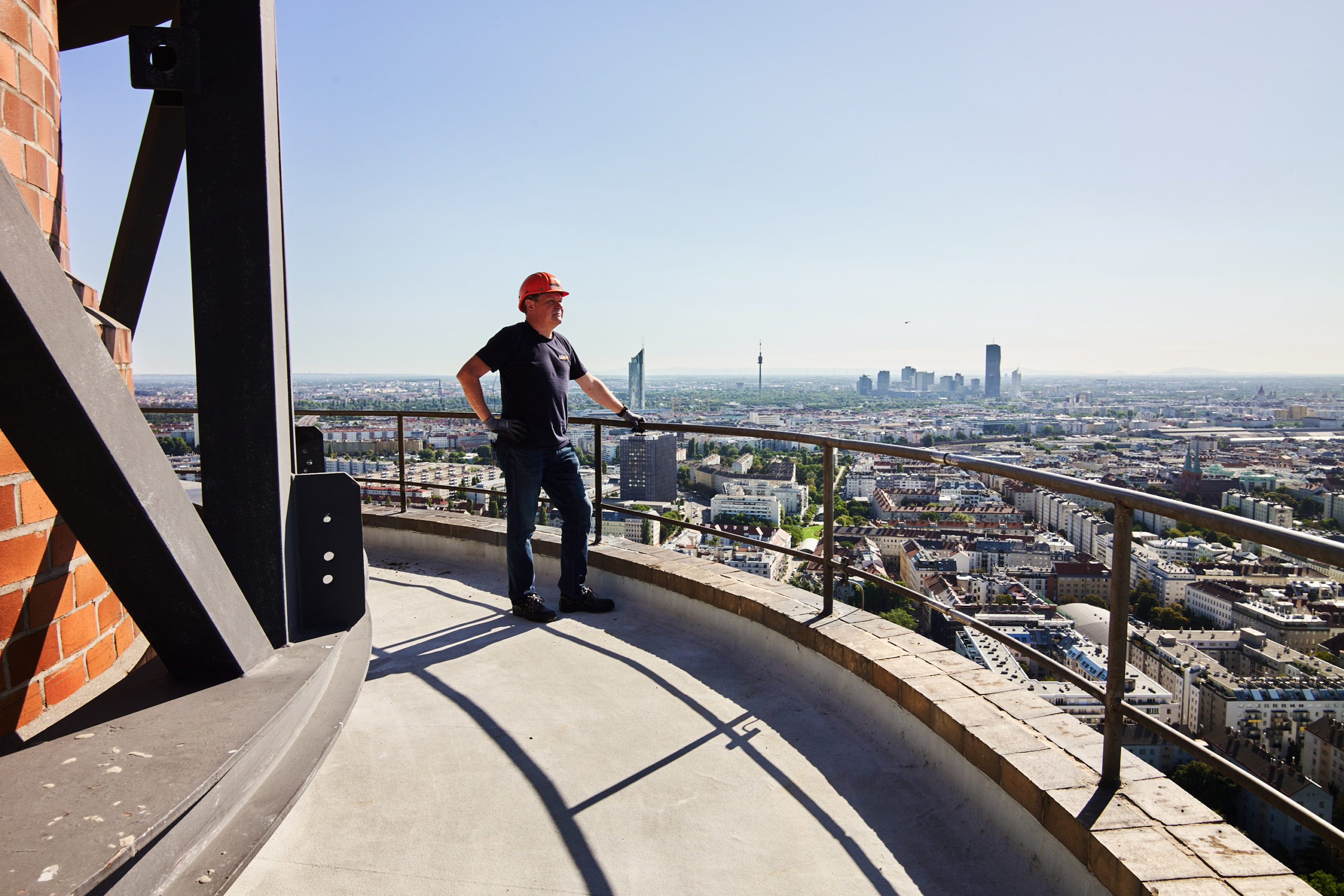 Ein Mann mit Schutzhelm steht auf einem hohen Balkon und überblickt die Skyline von Wien mit hohen Gebäuden in der Ferne.