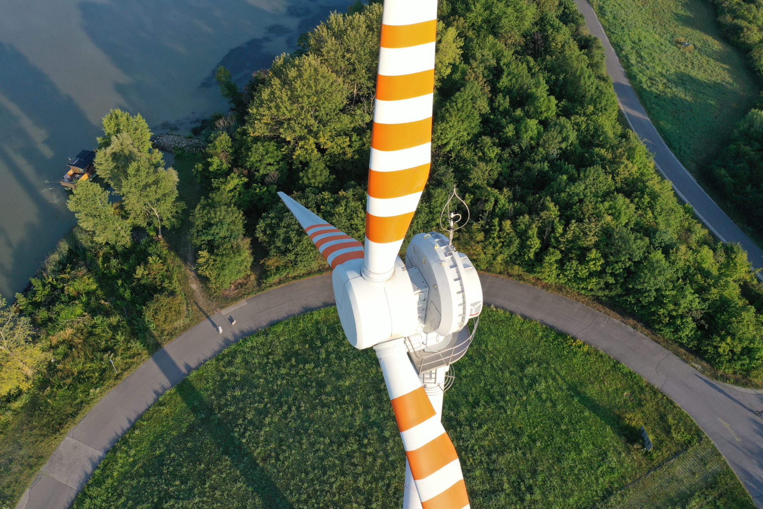 Draufsicht auf die Windkraftanlage Donauinsel mit orange gestreiften Rotorblättern, umgeben von Bäumen und einem geschwungenen Pfad in der Nähe eines Gewässers.