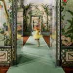 Eine verschwommene Gestalt in einem gelben Kleid dreht sich im lebhaften Bergl-Zimmer im Schloss Schönbrunn, dessen blumengeschmückter Flur mit grünem Teppich und verzierten Wänden eine verträumte Szene schafft.