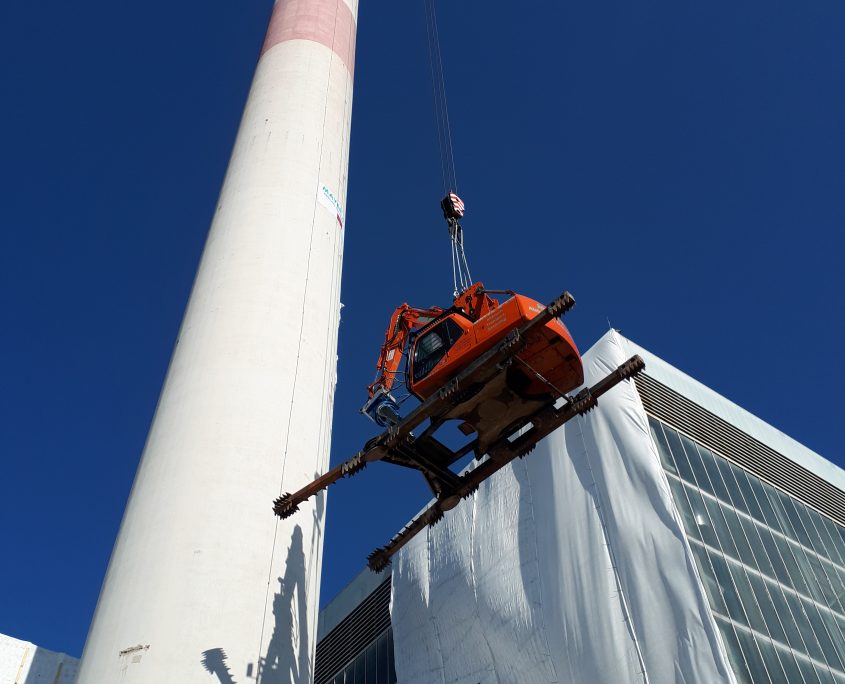 Ein Kran hebt ein orangefarbenen Bagger neben einem hohen Schornstein vor einem klaren blauen Himmel hoch.