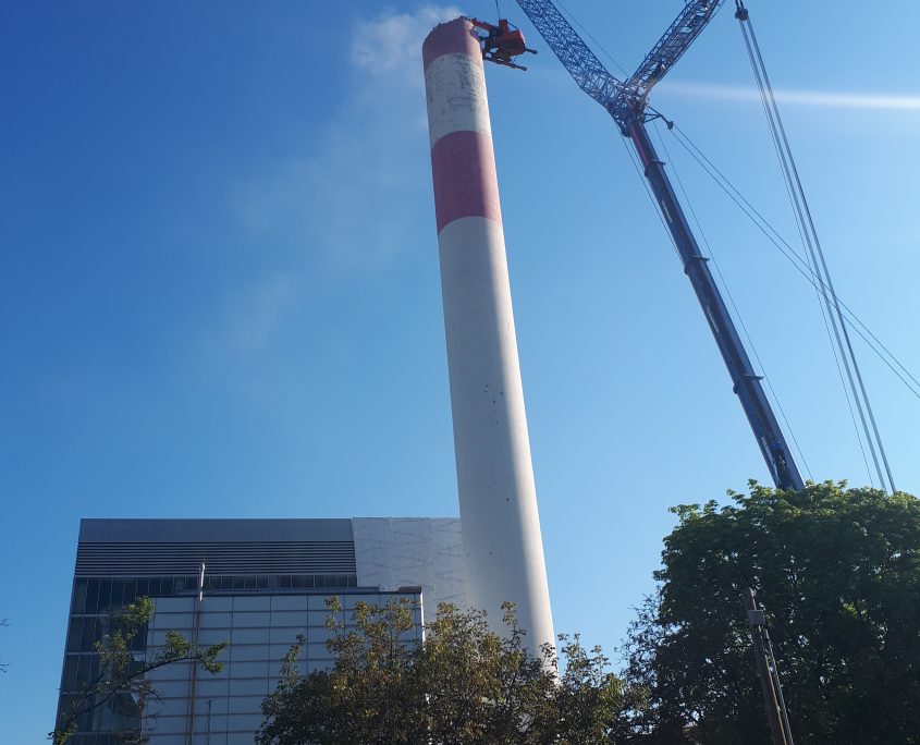 Ein Kran arbeitet unter einem klaren blauen Himmel an einem hohen Schornstein neben dem Fernheizwerk.