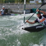 Zwei Kajakfahrer navigieren in einem Slalomkurs durch raues Wildwasser in der Vienna Watersports Arena auf der Donauinsel. Beide tragen Schutzausrüstung und sind hochkonzentriert.
