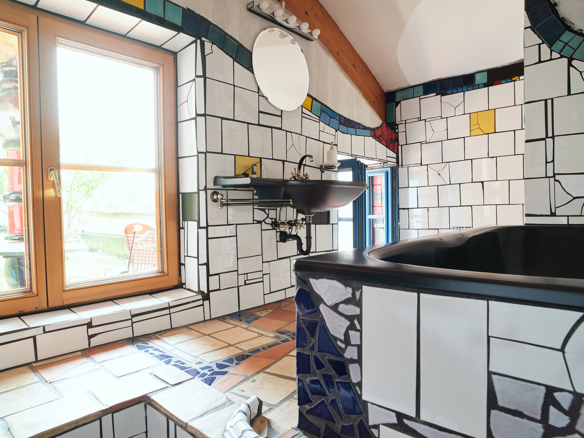 Badezimmer mit bunten Mosaikfliesen, rundem Spiegel, schwarzem Waschbecken und Badewanne. Großes Fenster sorgt für Tageslicht. Innenaufnahme des Kunsthaus Wien.