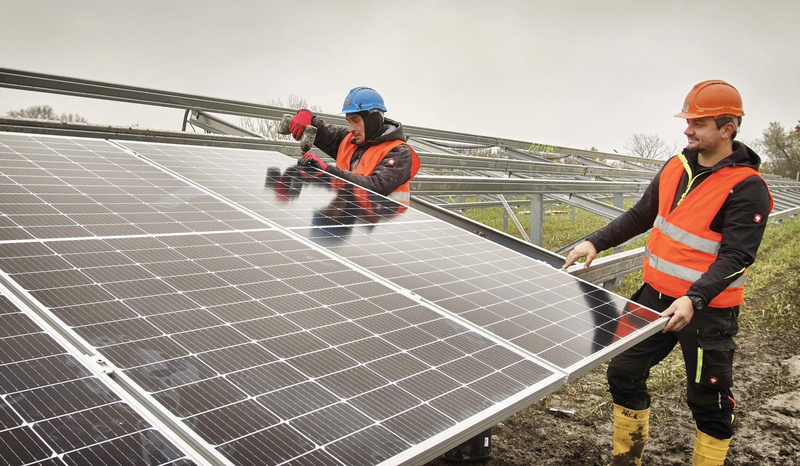 Zwei Arbeiter mit Helmen und Westen installieren an einem bewölkten Tag im Freien Solarmodule.
