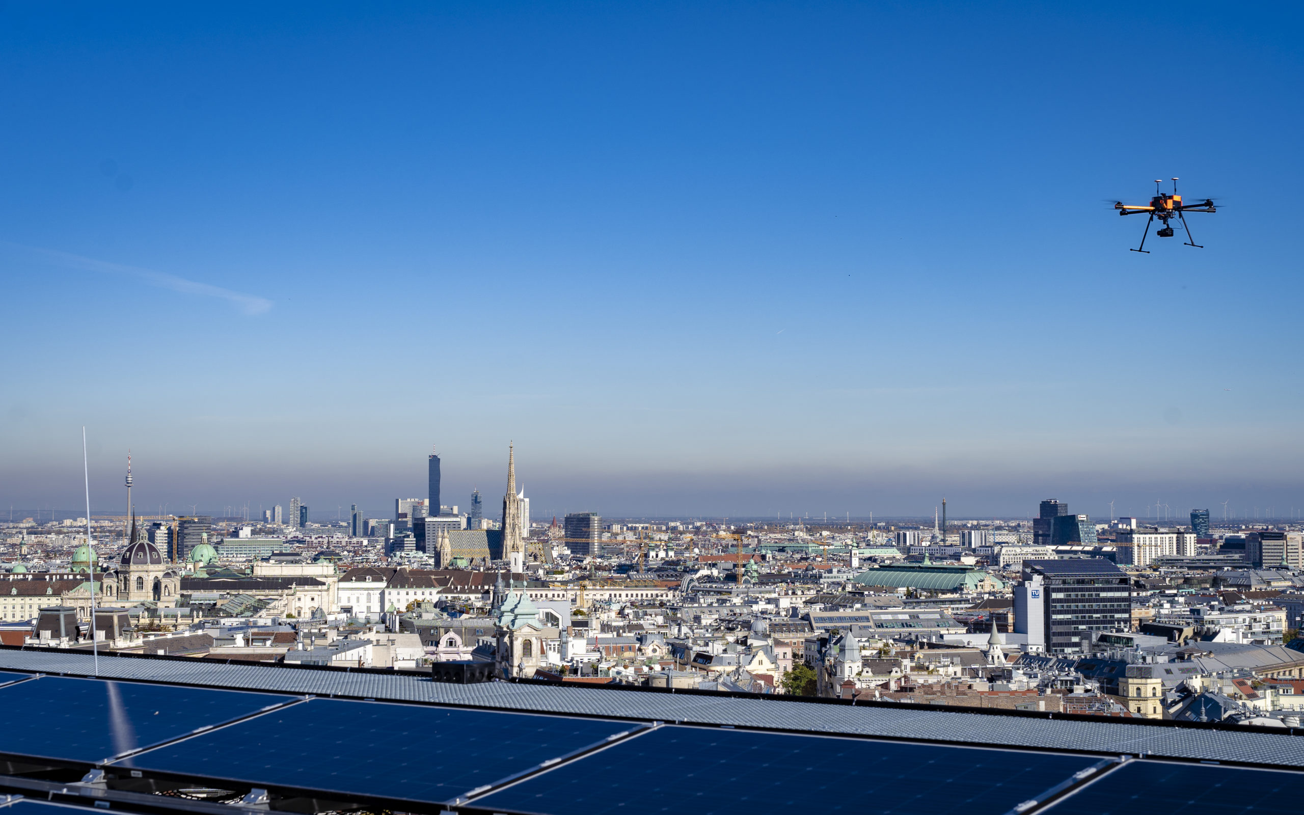 Drohne fliegt über die Wiener Stadtlandschaft. Im Vordergrund sind die Solarmodule des Haus des Meeres zu sehen, dahinter klarer, blauer Himmel.