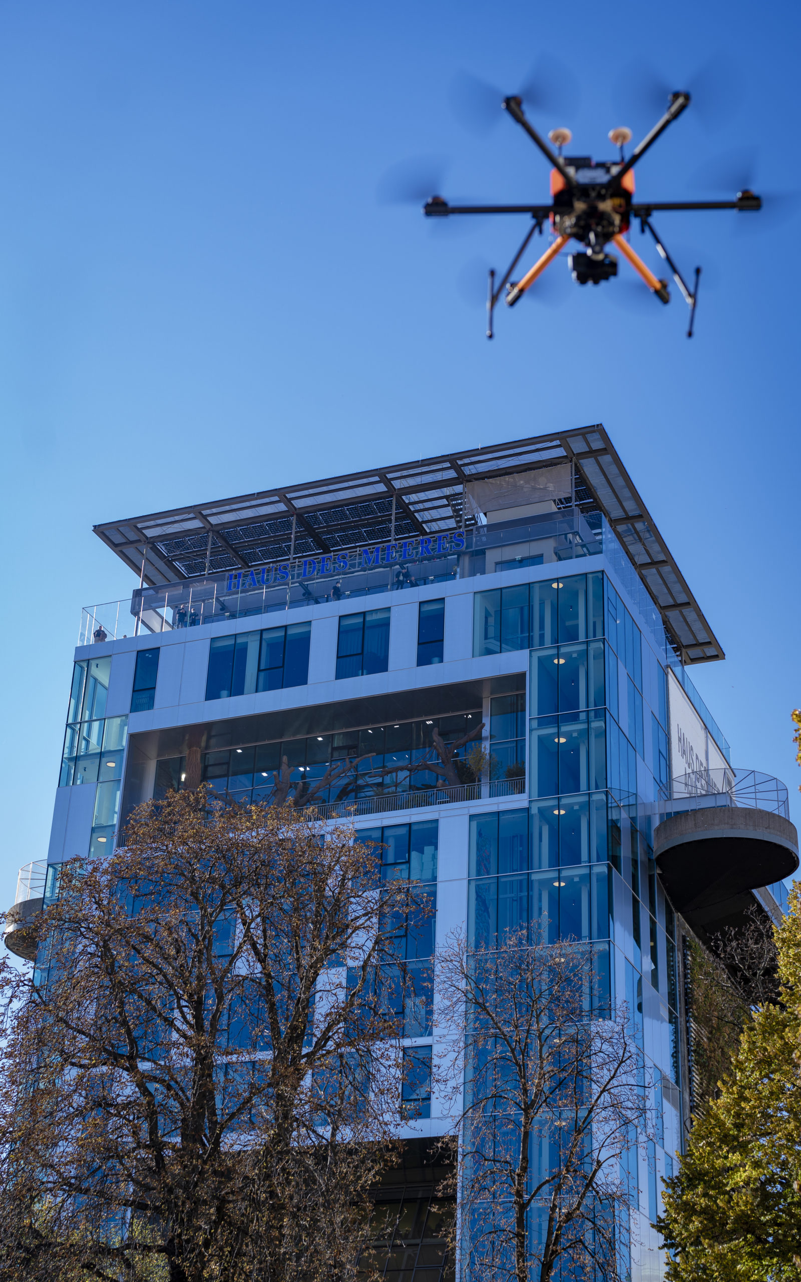 Vor dem klaren blauen Himmel gleitet eine Drohne anmutig an der modernen Glasfassade des Haus des Meeres vorbei, um die Photovoltaikanlage auf dem Dach zu warten.