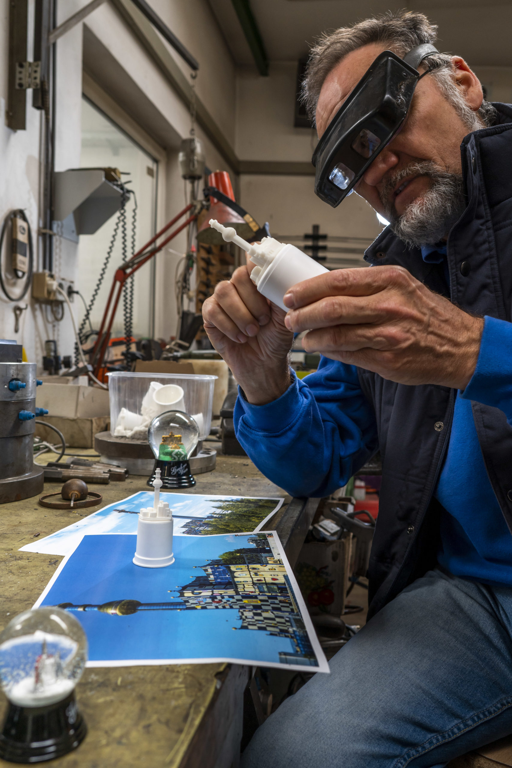 Eine Person mit Schutzbrille arbeitet in einer Werkstatt an einem Spittelau Modell, umgeben von Fotos und einer Schneekugel.