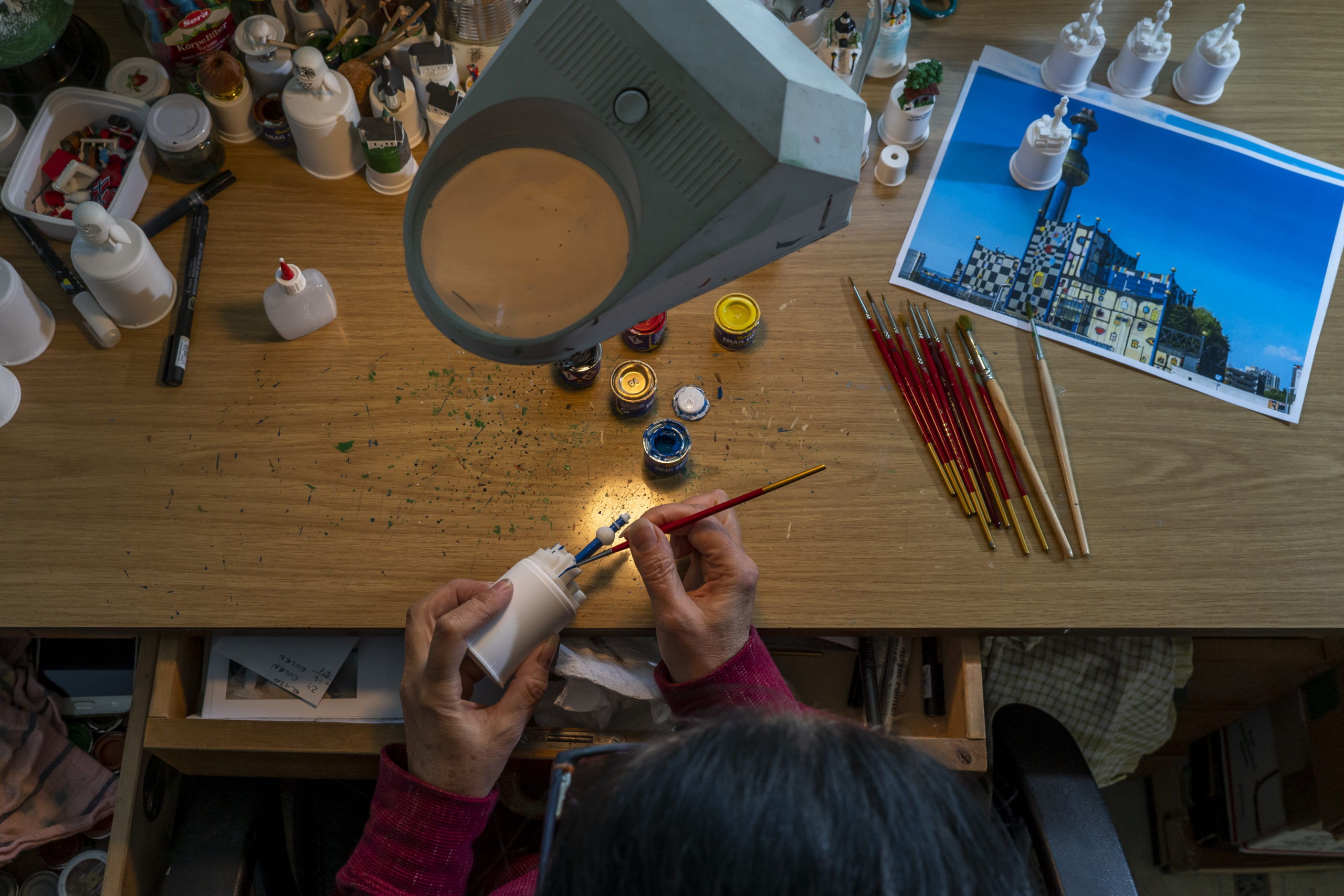 Eine Person bemalt unter einer Vergrößerungslampe mit Farben, Pinseln und einem Referenzfoto auf dem Tisch sorgfältig ein Modellgebäude.
